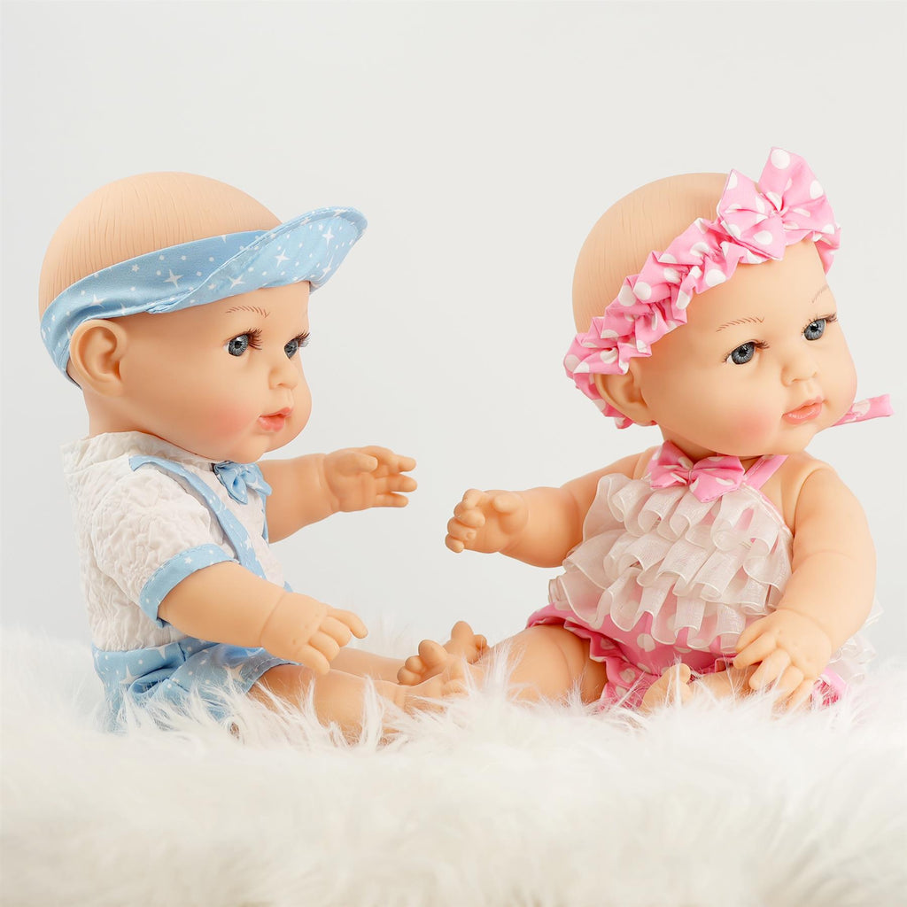 Two anatomically correct 14-inch twin dolls, one dressed in a blue outfit with a star-patterned visor and the other in a pink frilly outfit with a matching headband, are sitting on a soft white surface. The dolls have expressive faces and are posing as if interacting with each other. BiBi Dolls.