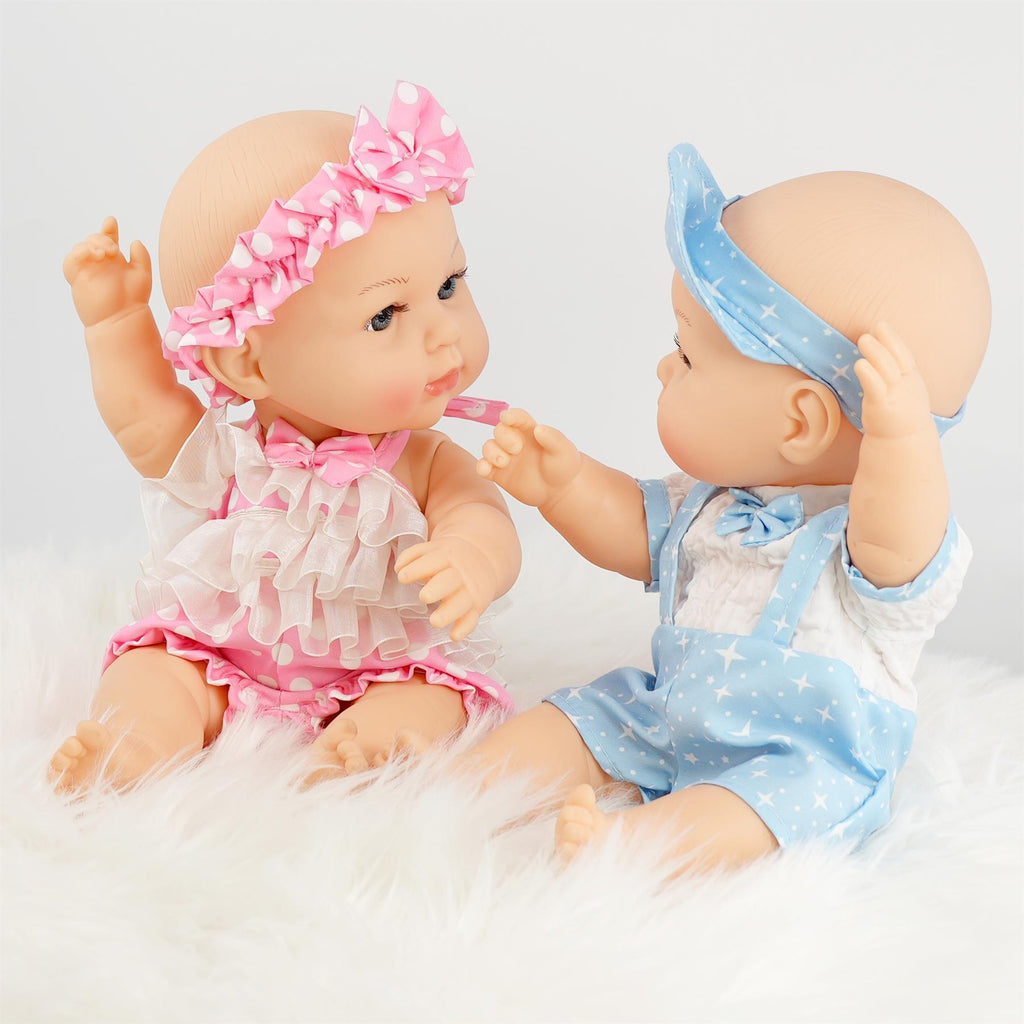 Two anatomically correct 14-inch twin dolls, one girl with a pink and white polka dot dress adorned with lace, and one boy dressed in a light blue outfit with a white shirt and star pattern. They are sitting on a fluffy white surface, engaging playfully. BiBi Dolls.