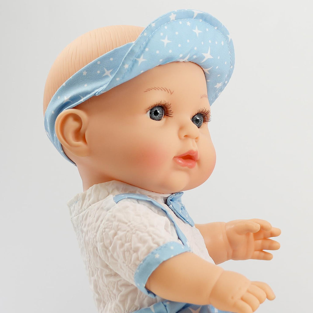 A close-up view of a 14-inch doll with light brown eyes and a soft, chubby face. The doll is wearing a white, textured shirt and blue straps, complemented by a blue visor adorned with white stars. This doll, designed to be anatomically correct, has a cheerful expression and is posed as if reaching out or playing. Perfect for imaginative play. BiBi Doll.