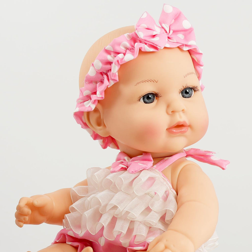 A close-up of a cute baby doll with a round head and open eyes, wearing a pink polka dot headband and a frilly white outfit. The doll has a soft expression and is posed sitting down, showcasing its adorable features. BiBi Dolls.